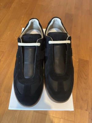 Maison Margiela Gats Svarta - Stilrena svarta sneakers från Maison Margiela med ovandel i mocka och läder, vita snören och klassisk gummisula. Snygg minimalistisk design med diskreta detaljer och präglad logga på plösen. Perfekta för dig som gillar exklusiva och tidlösa sneakers.