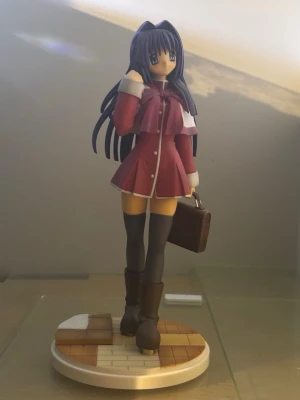 Anime figur cosplay skoluniform röd - Detaljerad animefigur i plast med lång mörkblå peruk, röd skoluniform med vita detaljer, höga stövlar och brun portfölj. Perfekt för cosplay eller maskerad. Står på en rund bas med golvmönster.