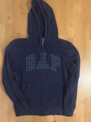 Mörkblå hoodie med dragkedja från GAP - Mörkblå hoodie från GAP med dragkedja framtill och stor huva. Klassisk GAP-logga i blått på bröstet och två fickor framtill. Tillverkad av 77% bomull och 23% återvunnen polyester för en mjuk och bekväm känsla. Perfekt för en avslappnad och stilren look.