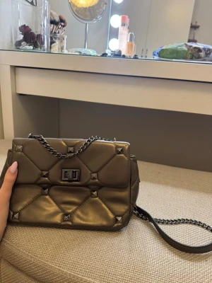 snygg quiltad metallic handväska - Cool quiltad handväska i metallic guld med kedjerem och dekorativa nitar. Väskan har ett vridlås framtill och fack inuti för bra organisering. Perfekt accessoar för att lyfta din outfit med definite warm vibes.