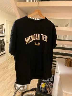 Svart Michigan Tech t-shirt - Snygg svart t-shirt med tryck 'MICHIGAN TECH' i vitt och gult på bröstet samt en liten logga under texten. Klassisk passform med rund hals och korta ärmar. Perfekt för dig som gillar college-vibe och enkel stil.