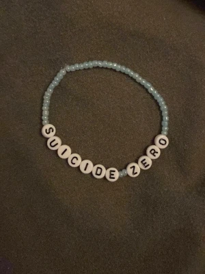 Pärlarmband Suicide Zero - Ett handgjort armband med ljusblå små pärlor och vita runda bokstavspärlor som bildar texten 'SUICIDE ZERO'. Perfekt accessoar för att visa stöd och sprida medvetenhet. Elastisk tråd för enkel användning.