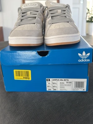 Grå Adidas sneakers med vita ränder - Snygga grå sneakers från Adidas med klassiska vita ränder på sidorna och stor logga på plösen. Ovandelen är i mocka och textil, med en vit sula och gummidetaljer. Perfekta för en stilren och sportig look. Helt oanvända, original förpackning skickas med,Pris kan diskuteras!