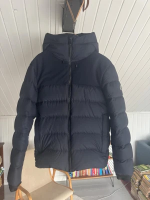 Mörkblå Sail Racing jacka - Snygg mörkblå Sail Racing pufferjacka med huva och dragkedja framtill. Jackan har breda, vadderade kanaler och en minimalistisk look. Storlek L i nyskick. Perfekt för kalla dagar och enkel att matcha med olika outfits. Justerbar huva och diskret logga på ärmen.