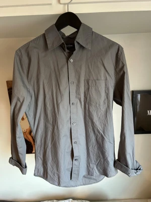 Grå skjorta med lång ärm - Stilren grå skjorta med klassisk krage och bröstficka. Skjortan har långa ärmar och knäppning framtill. Perfekt för en clean och enkel look. Passar dig som gillar minimalistisk stil.