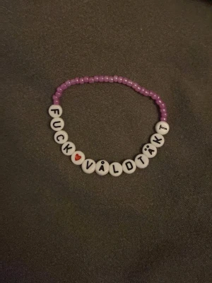 Lila pärlarmband med budskap - Ett handgjort armband med lila små pärlor och vita runda pärlor med svarta bokstäver som bildar texten 'FUCK VÅLDTÄKT'. Ett hjärta i rött finns också bland bokstäverna. Perfekt för dig som vill bära ett tydligt statement.