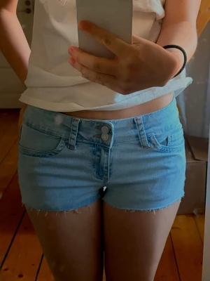 Ljusblå jeansshorts från Nelly - Ljusblå jeansshorts från Nelly med råa kanter och dubbla knappar framtill. Klassisk femficksmodell med bälteshällor och broderad detalj på bakfickorna. Perfekta för varma dagar och enkel att matcha med olika toppar.