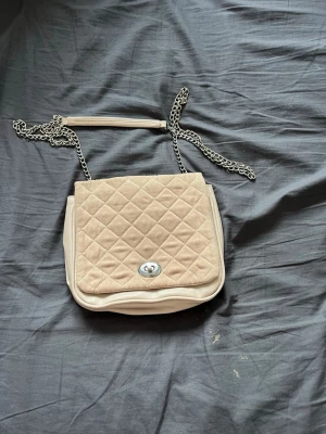 Beige quiltad axelremsväska med kedja - Snygg beige handväska med quiltat mönster på locket och silverfärgad kedjerem. Väskan har en klassisk rektangulär form och stängs med ett vridlås i metall. Materialet är en kombination av mockaimitation och slät konstläder.