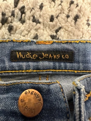 Ljusblå jeans från Nudie Jeans Co - Säljer ett par klassiska ljusblå jeans från Nudie Jeans Co med snygga kontrastsömmar och femficksmodell. Jeansen har en rak passform och är tillverkade i slitstark denim av bomull. Perfekta för en avslappnad och trendig look.