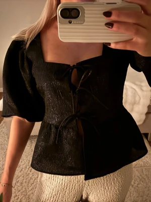 Svart blus med snörning och puffärm - Säljer en svart blus med glittrig finish, puffiga korta ärmar och snygg snörning framtill. Blusen har en fyrkantig ringning och är lätt figursydd för en modern look. Perfekt för dig som vill ha något trendigt och unikt.
