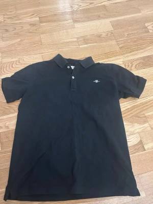 Svart pikétröja med liten logga - Klassisk svart pikétröja med krage och två knappar framtill. Liten broderad logga på bröstet. Perfekt till jeans eller shorts för en clean look.