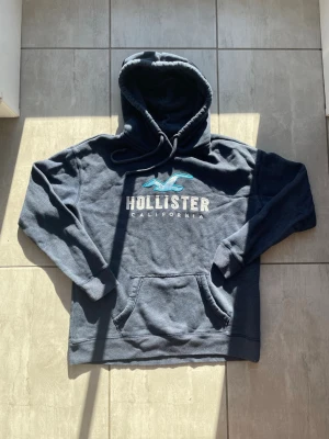 Mörkblå Hollister hoodie - Snygg mörkblå hoodie från Hollister med stor vit och turkos logga på bröstet. Klassisk känguruficka framtill och justerbar huva med dragsko. Mjuk och skön bomullsblandning, perfekt för chill dagar eller när du vill ha en avslappnad stil. Vid frågor eller funderingar skriv gärna! Passar Strl:M
