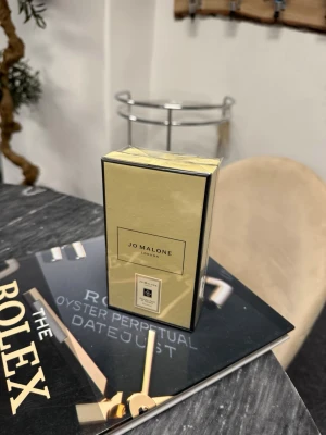 Jo Malone London parfym - Lyxig parfym från Jo Malone London i elegant guldfärgad och svart förpackning. Klassisk rektangulär kartong med minimalistisk design och tydlig logotyp framtill. Perfekt för dig som gillar exklusiva dofter och stilren estetik.