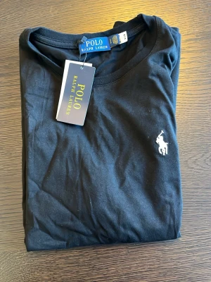 Svart t-shirt från Polo Ralph Lauren - Klassisk svart t-shirt från Polo Ralph Lauren med rund halsringning och ikoniska vita logobroderiet på bröstet. Tillverkad i mjuk bomull för en skön känsla och perfekt passform. En stilren basic som funkar till allt.