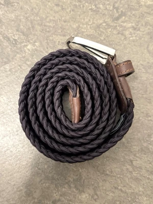 Blått flätat bälte från John Henric - Snyggt svart flätat bälte med bruna skinn-detaljer och silverspänne. Perfekt accessoar för att lyfta din outfit med en stilren touch. Bältet är från John Henric och är använt endast ett fåtal gånger. Nypris: 499kr.