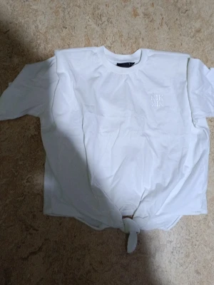 Vit topp med knyt från NIK&NIK - Snygg vit topp från NIK&NIK med knyt framtill. Toppen har rund halsringning och korta ärmar. Diskret logga på bröstet ger en clean look. Perfekt att matcha med jeans eller kjol för en trendig stil.