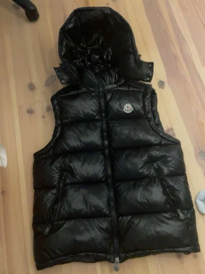 Svart dunväst från Moncler med huva - Snygg svart dunväst från Moncler med glansig finish och avtagbar huva. Västen har två dragkedjeförsedda fickor framtill och Moncler-logga på bröstet. Perfekt för lager-på-lager och håller dig varm under kyliga dagar.