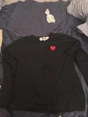 Svart sweatshirt med rött hjärta - Svart sweatshirt från PLAY Comme des Garçons med det ikoniska röda hjärtat med ögon broderat på bröstet. Klassisk rund halsringning och lång ärm. Perfekt för en stilren och trendig look.