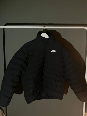 Svart pufferjacka från Nike - Säljer en svart pufferjacka från Nike med hög krage och dragkedja framtill. Jackan har en klassisk pufferdesign med horisontella sömmar och Nike-logga i vitt på bröstet. Perfekt för kalla dagar och riktigt snygg till streetwear-stilen. Barnstorlek men stämmer överens med en XS