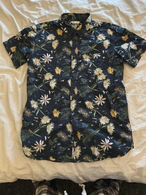 Blommig kortärmad skjorta, barn XS.  - Snygg kortärmad skjorta i mörkblått med blommigt mönster i vitt, gult och grönt. Skjortan har klassisk krage, knappar framtill och är perfekt för en chill och stilren look. Tillverkad i mjuk bomull som känns skön mot huden.