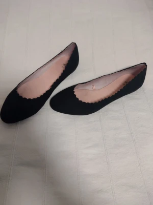 Svarta ballerinaskor med vågig kant - Säljer ett par svarta ballerinaskor från H&M med snygg vågig kant runt öppningen. Skorna har en klassisk rundad tå och är gjorda i ett mjukt tygmaterial med ljusrosa innersula. Perfekta för en stilren och enkel look. Har bara provats men det rosa tyget invändigt är lite gråflammigt.