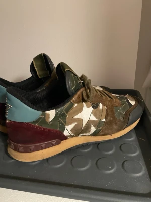 Valentino rockrunners  - Snygga sneakers från Valentino med camomönster ir grönt, beige och svart. Skorna har bruna mockadetaljer, blå och vinröd häl samt coola nitar bak. Yttermaterialet är en mix av textil, mocka och läder. Perfekta för dig som vill sticka ut med din stil.