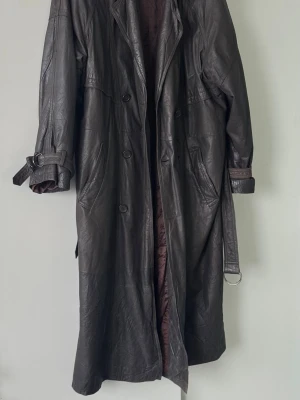 Lång svart skinnjacka med bälte - Snygg lång svart skinnjacka med bälte i midjan och vid ärmsluten. Jackan har bred krage, knäppning framtill och två stora fickor. Perfekt för dig som vill ha en cool och tidlös look.