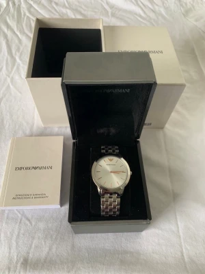 Silverfärgad klocka från Emporio Armani - Stilren klocka från Emporio Armani med silverfärgat metallarmband och vit urtavla. Det är en original klocka som aldrig har används, bara stått och samlat damm. Den vill bäras av någon som har klass💫💫OBS: batteri måste bytas