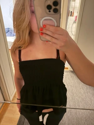 Svart volang linne med tunna band - Denna tröja e helt oanvänd och i ett mycket bra skick. Modellen är figurnära upptill och har en lätt utställd nederdel som ger en söt volang-effekt. Perfekt till jeans eller kjol för en snygg look. Materialet känns mjukt och stretchigt.