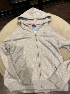Grå zip hoodie från Gant - Snygg ljusgrå hoodie från Gant med dragkedja framtill och huva. Broderad Gant-logga på bröstet och texten 'Good to Great' på andra sidan. Mjuk bomullsblandning och ribbade muddar. Insidan av huvan har en röd, vit och marinblå rand för extra detalj. Det finns några små fläckar så dm för bilder på dom men annars så Pris går att diskuteras och dm för mer bilder och info! 