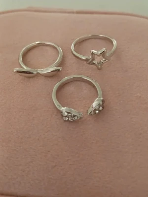 Söta silvriga ringar med stjärna & hjärta💕💕 - Tre silvriga ringar i olika designer: en med två vingar en med en stjärna och en öppen ring med två hjärtan dekorerade med små glittrande stenar. Perfekta för att mixa och matcha till din stil. Aldrig använt.