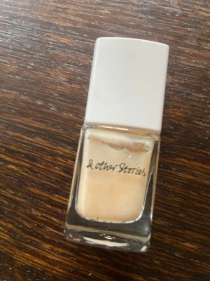 Pärlemorskimrande nagellack Seashell - Nagellack från & Other Stories i nyansen Seashell. Färgen är ljus pärlemor med subtilt skimmer, perfekt för en fräsch och elegant look. Kommer i en stilren fyrkantig glasflaska med vit kork. Lacket är lätt att applicera och ger en snygg finish.