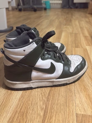 Nike Dunk High svart/vit sneaker - Nike Dunk High sneakers i svart och vitt läder med klassisk Swoosh på sidan. Skorna har högt skaft, svart snörning och perforerad tåbox för extra ventilation. Fodret är svart och sulan är vit med svart yttersula. Perfekt för dig som gillar retrostil.