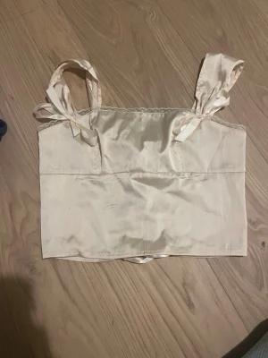 Beige satin linne med korsett rygg och knytband för axlarna - Säljer detta för att det inte passar. Snyggt beige linne i satin med axelband som knyts på axlarna. Baksidan har en snörning med satin band. Den är i storlek S men passar S-M pga snörningen där bak. Inga defekter. Priset kan diskuteras 💕