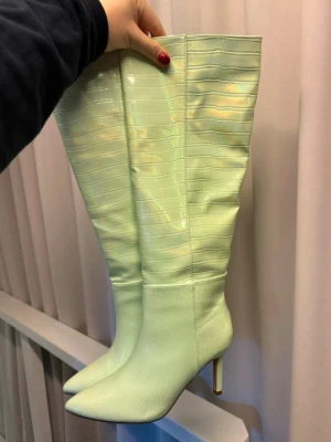 Ljusgröna höga boots med krokopräglad yta - Säljer ett par höga boots i limegröna med spetsig tå och smal klack. Stövlarna har en glansig, krokopräglad yta som ger en trendig look. Perfekta för dig som vill sticka ut med färg och mönster. Materialet ser ut som konstläder. Stövlarna har utgått från gina tricot 