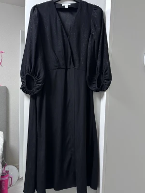 Svart långklänning med puffärm - Svart långklänning med v-ringning och vida puffärmar. Klänningen har en lätt glittrig yta och är sydd i ett mjukt, fallande tyg. Perfekt för dig som gillar en stilren look med det lilla extra.