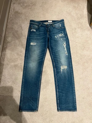 Blå slitna jeans från tiger of swedan - Säljer ett par blå jeans från tiger of swedan med snygga slitningar och distressed detaljer på framsidan. Klassisk femficksmodell med raka ben och läderpatch bak i midjan. Perfekta för en avslappnad och trendig look. Tillverkade i bomull.