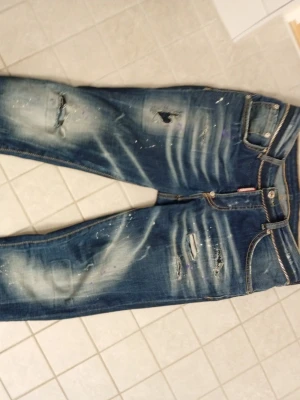 Blå slitna jeans från Dsquared2 - Säljer ett par blå jeans från Dsquared2 med slitningar, hål och färgstänk för en riktigt edgy look. Jeansen har klassisk femficksmodell, dragkedjegylf och kontrastsömmar. Perfekta för dig som gillar streetstyle och vill sticka ut.