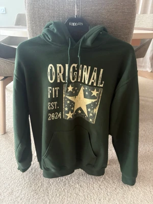 Mörkgrön hoodie med tryck från KappAhl - Mörkgrön hoodie från KappAhl med stor ficka framtill och huva med dragsko. Tryck på bröstet med texten 'ORIGINAL FIT EST. 2024' och en stor stjärna med flaggdetaljer. Mjuk och skön, perfekt till chill dagar.