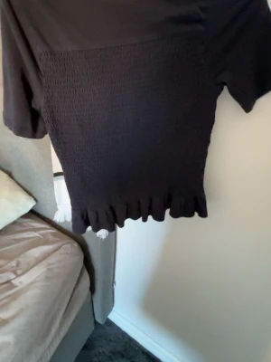 Svart croppad topp med volangkant - Svart croppad topp med korta ärmar och smockad framsida. Toppen har en söt volangkant nertill och är perfekt för en trendig look. Materialet känns mjukt och stretchigt, vilket gör den bekväm att bära.