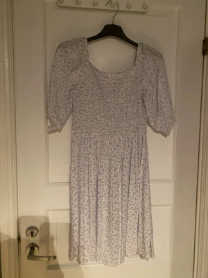 Ljusblå blommig kortklänning - Söt kortklänning med ljusblått blommönster och smockad överdel. Klänningen har puffiga kortärmar och en fyrkantig urringning. Perfekt för sommarens varma dagar och har en luftig, flowy kjol. Materialet känns som bomull eller polyester.