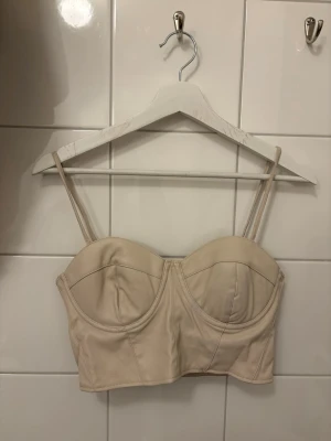 Beige korsett-topp med smala axelband - Snygg beige korsett-topp med smala axelband och markerade kupor. Toppen har dragkedja i ryggen och en croppad passform som ger en trendig look. Perfekt att styla med jeans eller kjol för en cool vibe.