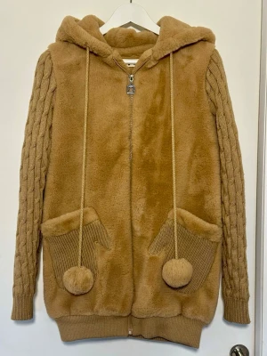 Beige teddyjacka med stickade ärmar - Mysig beige teddyjacka med huva, dragkedja och stora fickor framtill. Ärmarna är stickade med flätmönster och jackan har långa snören med bollar. På ryggen finns broderad text och ett pappersflygplan. Perfekt för dig som gillar en chill och trendig stil.