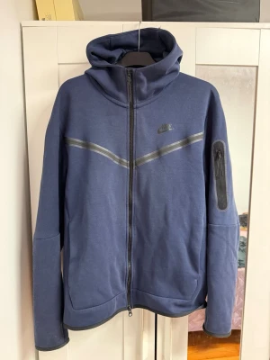 Blå Nike hoodie med dragkedja - Snygg blå hoodie från Nike med hel dragkedja, huva och gråa detaljer över bröstet. Har en praktisk ficka med dragkedja på ärmen och två fickor framtill. Perfekt för en sportig och avslappnad stil.
