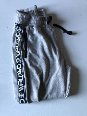 Grå mjukisbyxor World VRLWD - Grå mjukisbyxor med svart-vita detaljer från H&MD. Byxorna har resår och dragsko i midjan, svarta dragkedjor vid fickorna och en svartvit logga längs sidorna. Perfekta för en avslappnad och sportig stil.