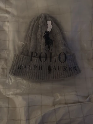 Grå Polo Ralph Lauren mössa - Snygg grå Polo Ralph Lauren mössa med uppvikt kant och klassisk logga. Tillverkad i mjukt material som håller dig varm och stilsäker under kalla dagar. Perfekt accessoar för en trendig och avslappnad look.