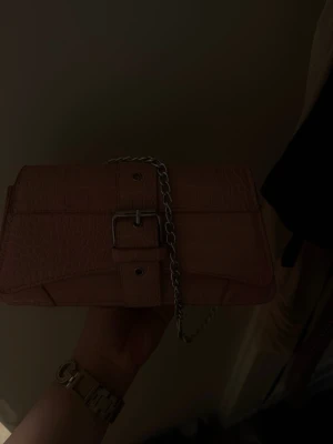 Babyrosa väska med kedja - Snygg rosa handväska/ axelväska i fejkläder med croco-mönster. Väskan har ett dekorativt spänne framtill och en silverfärgad kedja samt ett längre band till