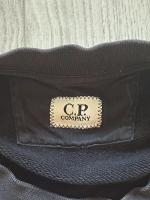 Svart sweatshirt från C.P. Company - Snygg svart sweatshirt från C.P. Company med rund halsringning och diskret logga på bröstet. Tröjan har ribbade muddar vid ärmslut och nederkant. Perfekt för en stilren och avslappnad look.