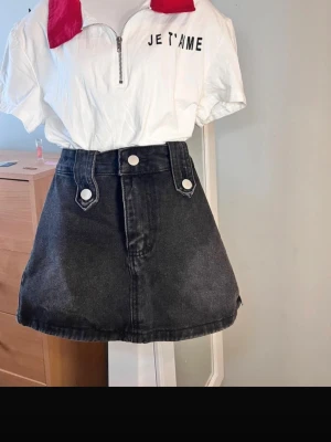 Set 2st Svart jeanskjol med knappar och t shirt  - Snygg svart kort jeanskjol med hög midja och dekorativa vita knappar framtill. Kjolen har breda bälteshällor och en klassisk A-linjeform som ger en trendig siluett. Perfekt att matcha med en t-shirt eller hoodie.