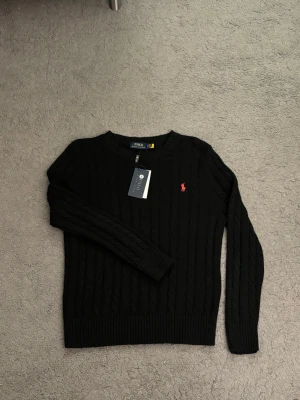 Svart kabelstickad tröja Polo Ralph Lauren - Svart kabelstickad tröja från Polo Ralph Lauren med rund halsringning och röd broderad logga på bröstet. Tröjan har långa ärmar och ribbade muddar vid ärmslut och nederkant. Perfekt för en stilren och klassisk look.